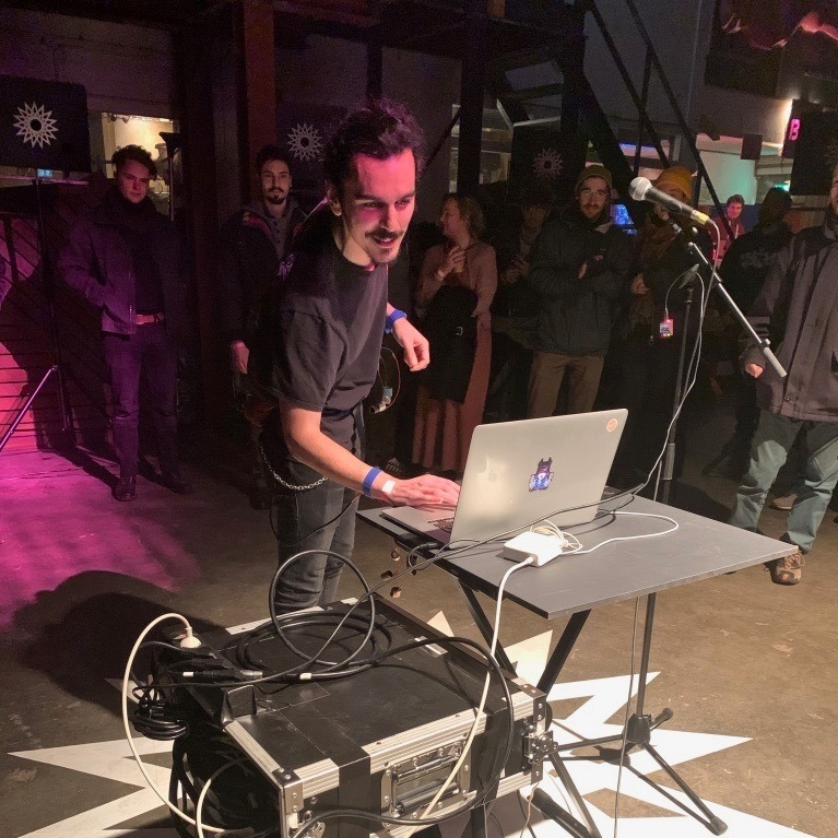 Görkem Arıkan is standing behind a table with a computer sitting on it. Multiple electronic devices are connected to the computer. His hand lays on the computer. He is seemingly amidst a performance. Around him and in the background are an audience of people standing and observing.