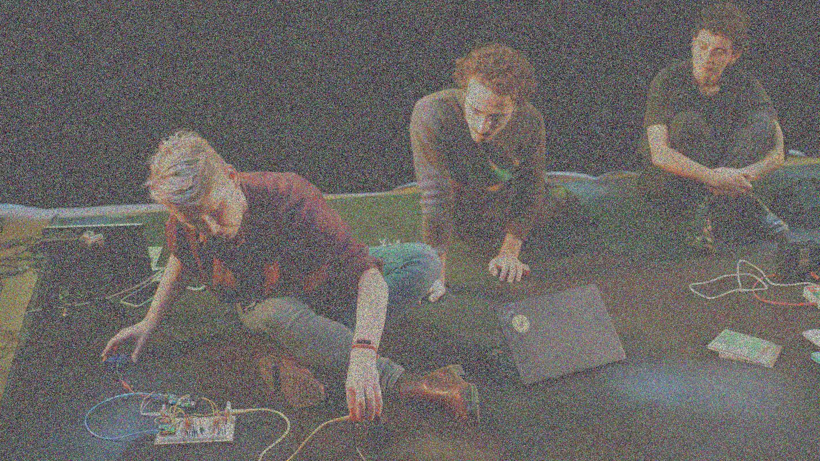 Drie mensen zitten op de vloer en kijken naar de persoon die elektronische prototypes met veel draden manipuleert.