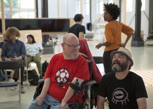 Speakers Vasilis Van Gemert and Eric Groot Kormelink are both smiling. Eric is in a wheelchair wearing a red T-shirt with his face turned towards Vasilis. Vasilis has a beard and is wearing a black T-shirt and a straw hat. He is in a squatting pose.