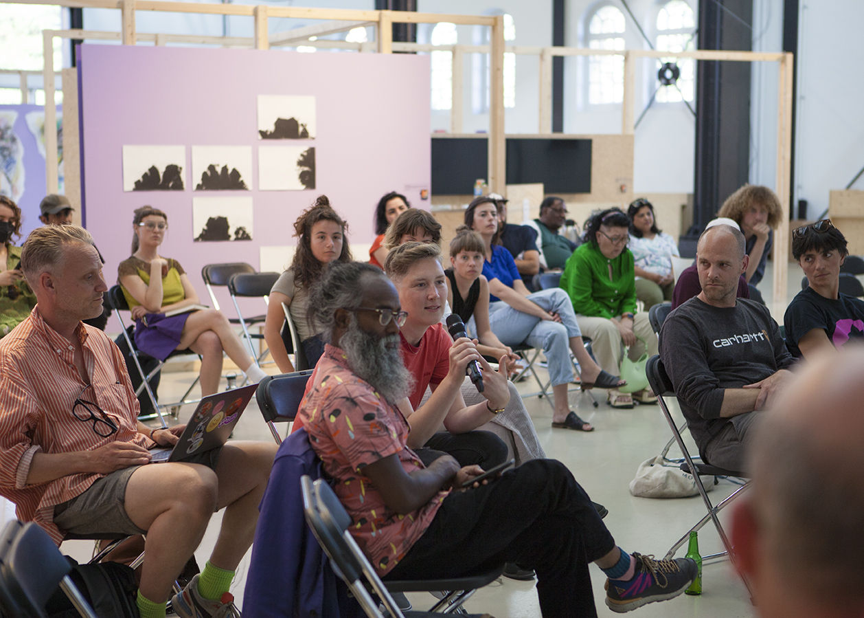 A large bright room in an exhibitions space holds space for an audience of about 20-25 people. Hackers and designers member Loes, is part of the audience and is asking a question via a wireless microphone.