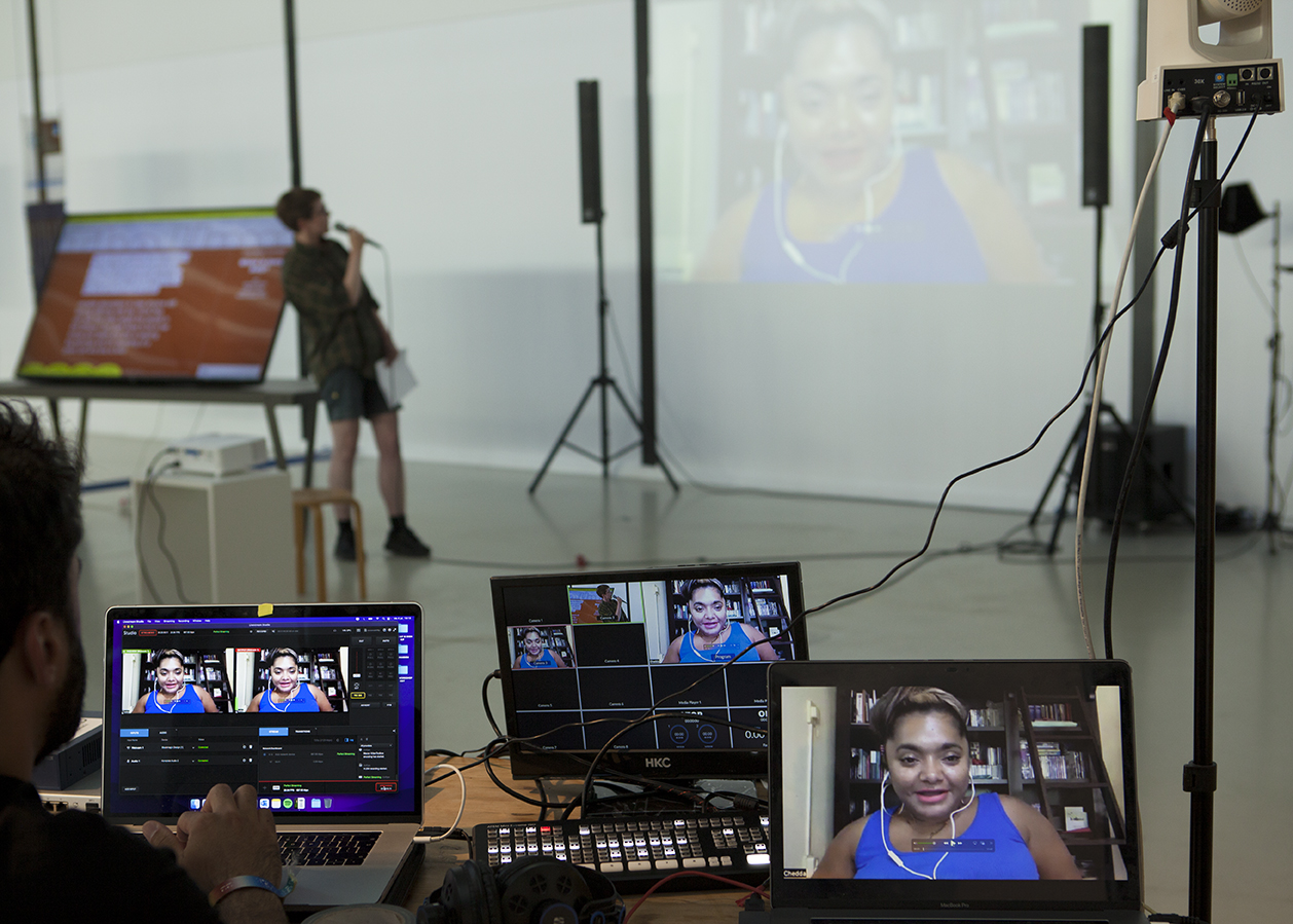 In the foreground are three computer monitors working to stream online. Cables run diagonally across the image connecting screens to camera. A blurry background shows speaker Jeanette Chedda being introduced by a person holding a microphone. A video of Jeanette’s face is projected on the wall and the same image is shown on the screens. Jeanette’s face is captured mid-sentence while wearing a clear blue sleeveless top and white headphones.