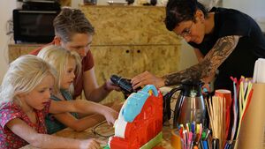 Twee workshopbegeleiders helpen twee workshopdeelnemers die aan een tafel zitten bij het gebruik van een boormachine op een felgekleurd plastic speelgoed.