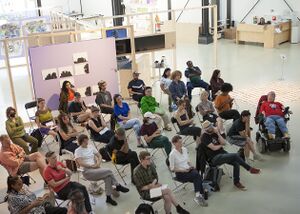 An audience captured from above. About 20-25 people seems focuced with attention towards a stage that is not featured on the image. The room is bright with high celings and has several artworks on display.