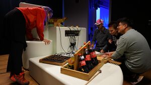 Workshopdeelnemers van verschillende leeftijden stemmen en spelen met de speciaal gebouwde instrumenten van SoundLAB in een geluidsgeïsoleerde glazen ruimte.
